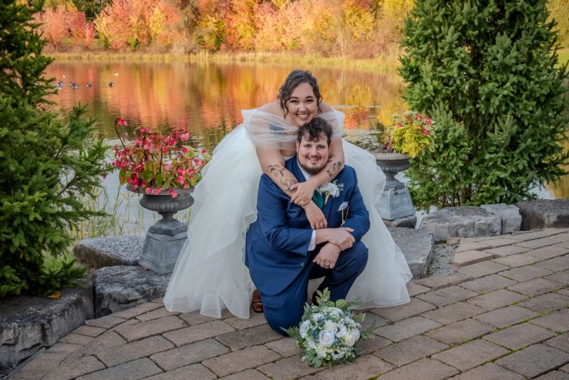 A bride perched on her groom's shoulders by a pond, both dressed in wedding attire, bathed in autumn's golden light.