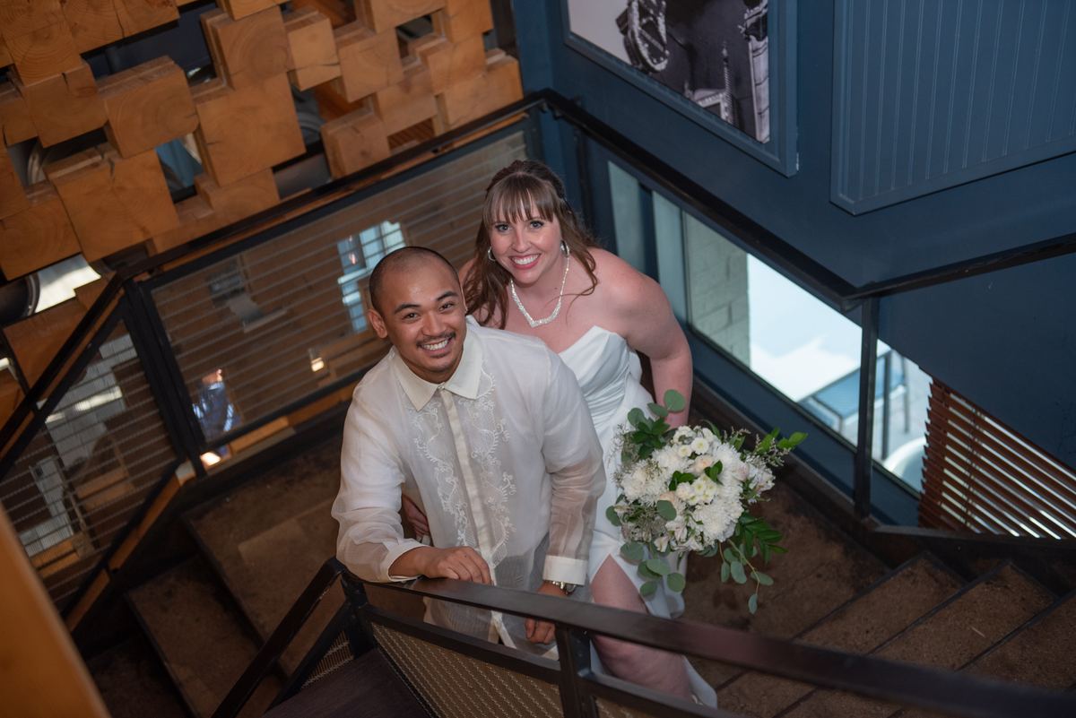 A smiling bride and groom stand on a modern wooden staircase, surrounded by warm lighting and geometric architecture.