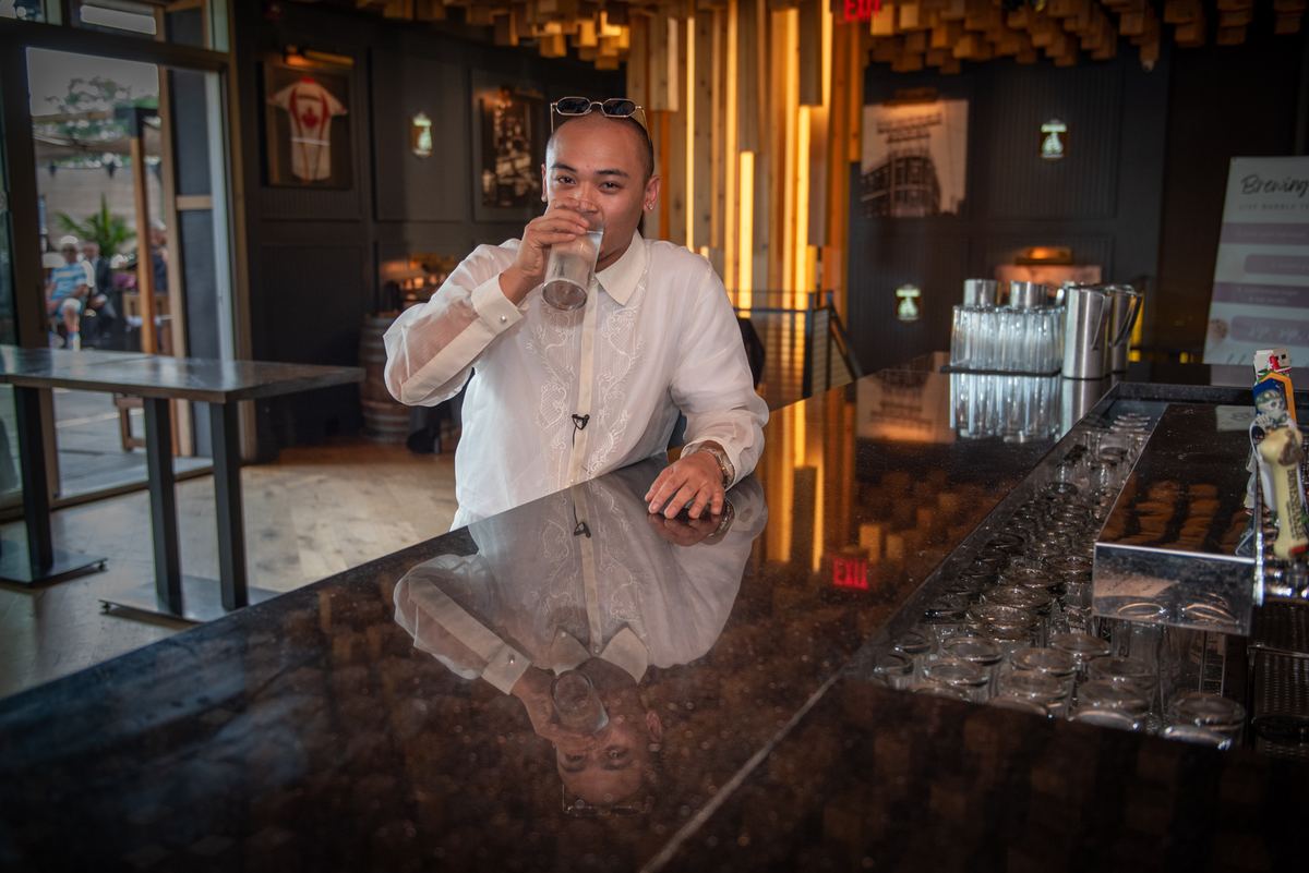 A man in a white embroidered barong sips from a glass at a glossy black bar, reflecting the surroundings.