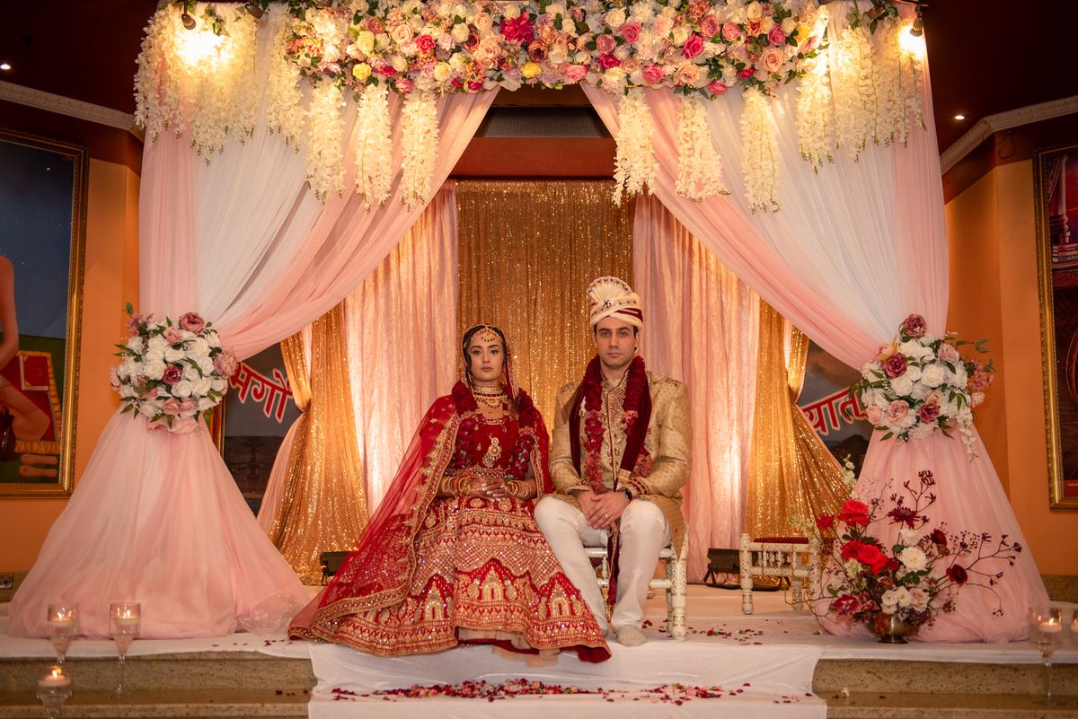 A newlywed couple in traditional attire sits on a decorated stage under a floral canopy, illuminated by warm lighting.