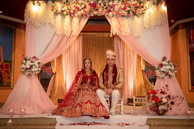 A newlywed couple in traditional attire sits on a decorated stage under a floral canopy, illuminated by warm lighting.