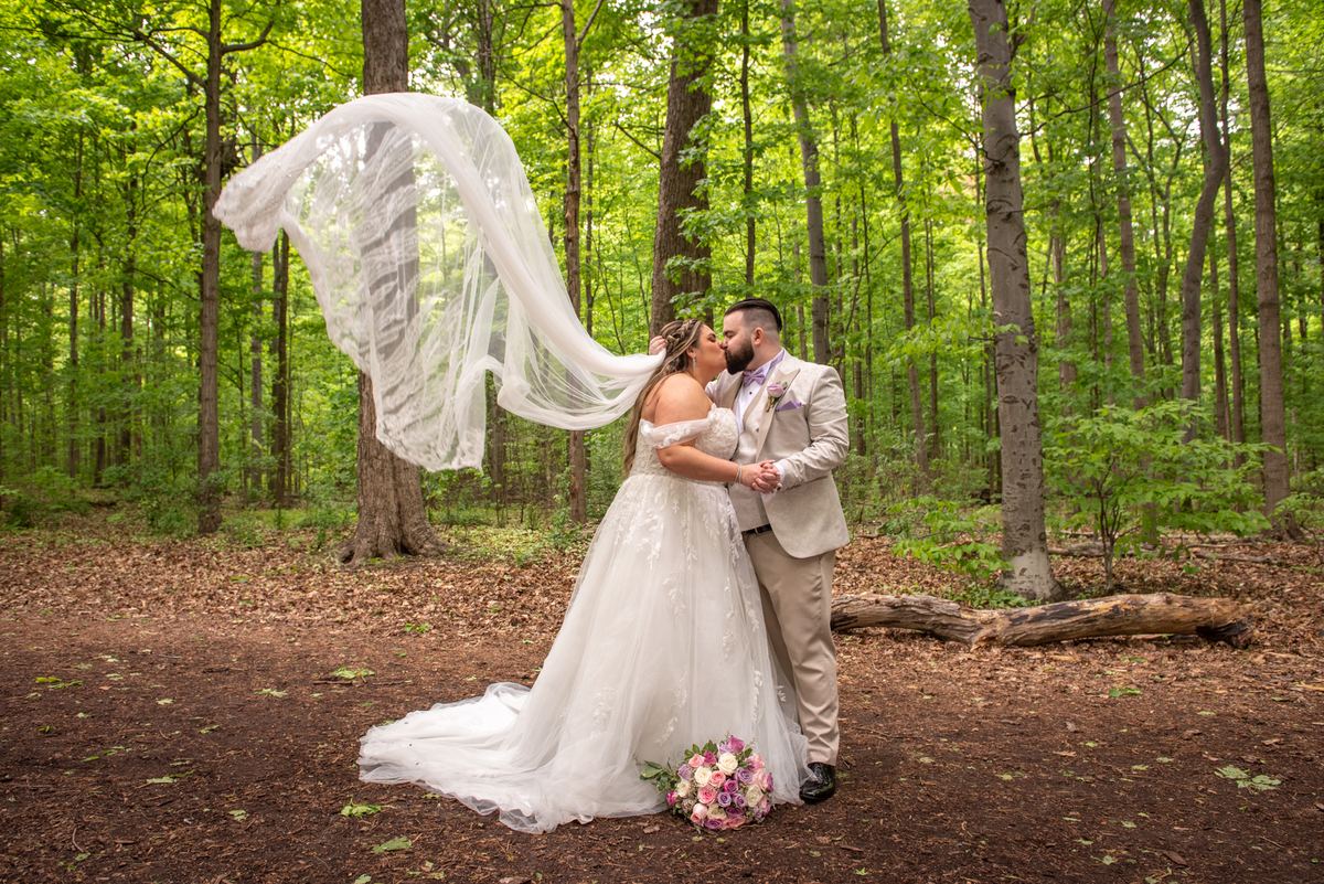 A bride and groom kiss in a sun-dappled forest, their white gown and light suit contrasting with the earthy floor.