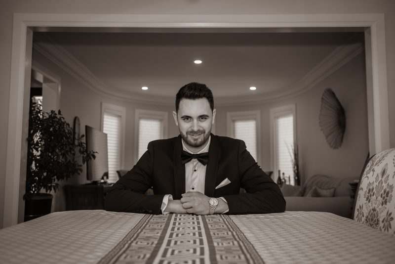 A man in a tuxedo smiles at the camera in a monochrome room with elegant curved architecture and recessed lighting.