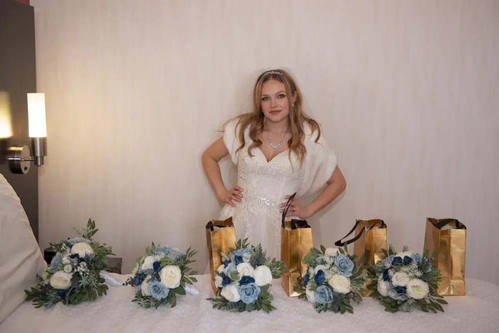 Nicole stands in her wedding dress behind blue and white bouquets and gold gift bags in the hotel room.