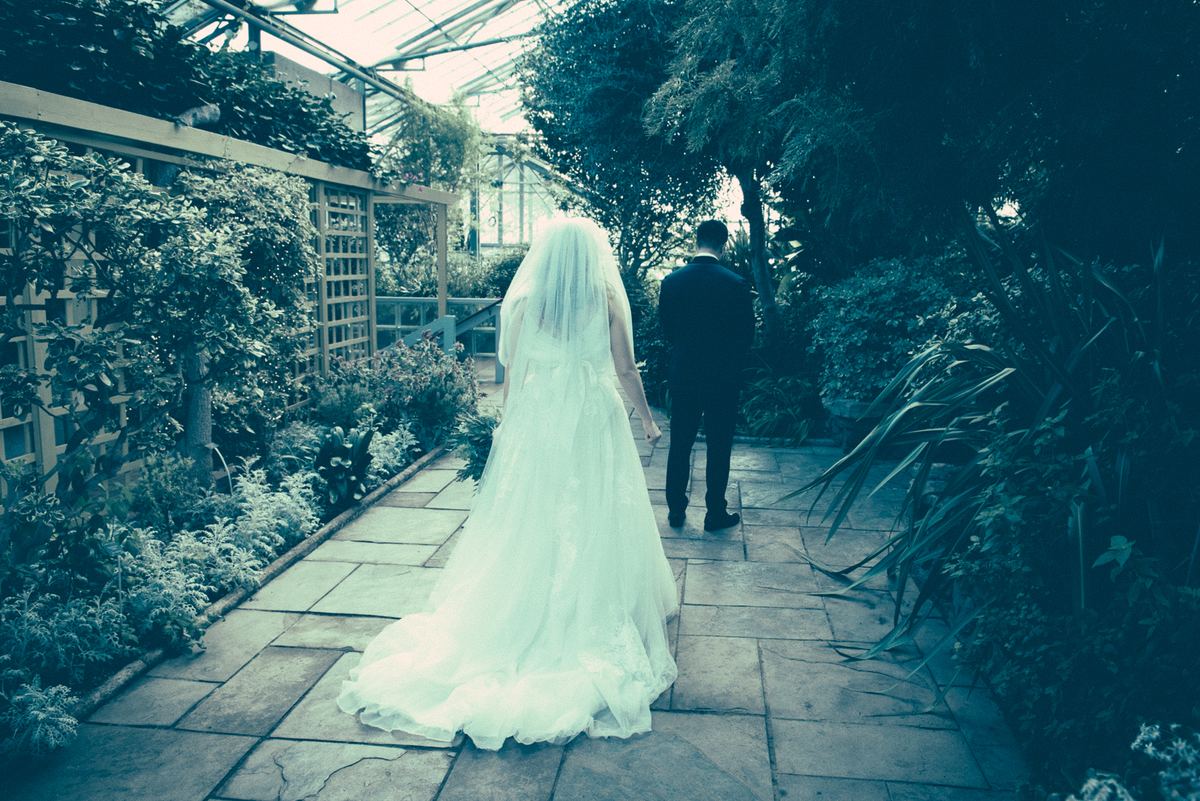 A bride and groom walk down a stone path in a lush greenhouse garden, backlit by diffused light through the glass ceiling.