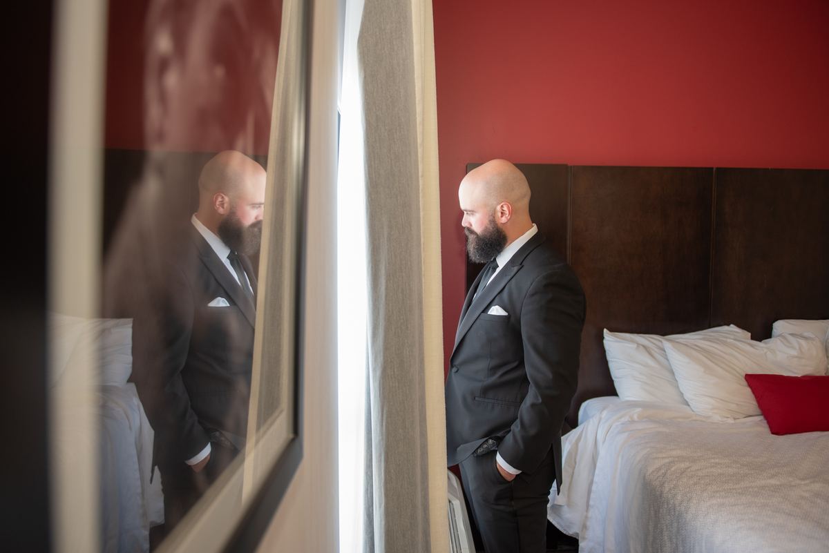 A man in a black suit stands by a window, gazing thoughtfully outside, with a red wall and white bedding visible behind him.