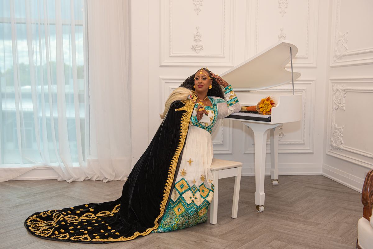 A woman in an ornate gold-embroidered gown leans beside a white grand piano in a bright room with soft daylight.