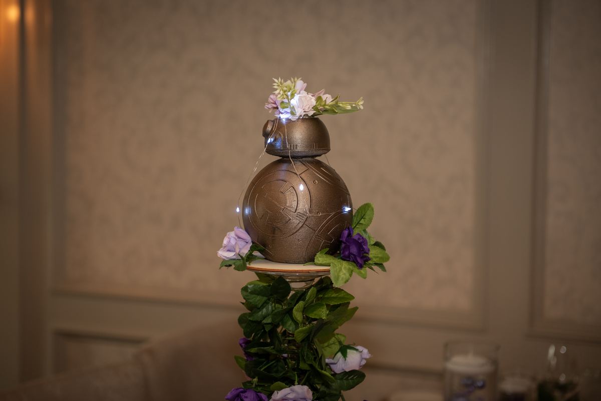 A decorated BB-8 droid with flowers and string lights stands on a floral platform, illuminated by soft ambient lighting.