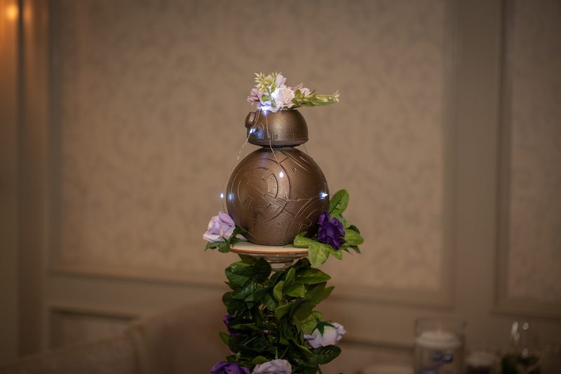 A decorated BB-8 droid with flowers and string lights stands on a floral platform, illuminated by soft ambient lighting.