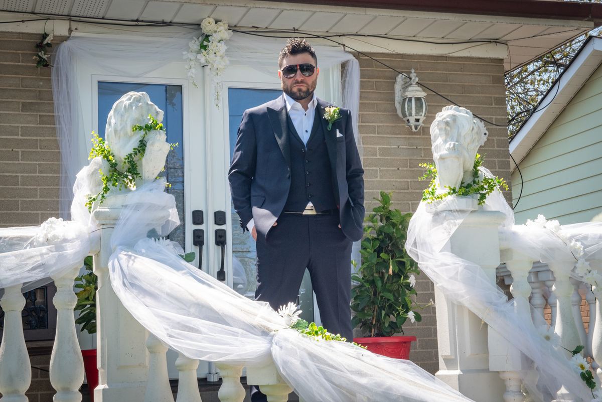 A groom in a dark suit and sunglasses stands confidently on a decorated porch with floral arrangements.