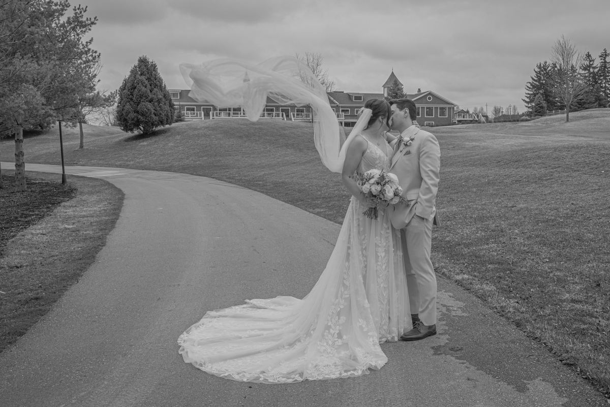 A bride and groom kiss passionately on a windy path, surrounded by trees and a large house, in black-and-white.