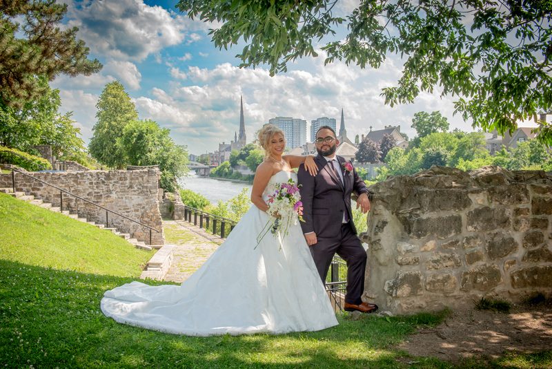 A smiling bride in a white gown and groom in a dark suit pose beside a stone wall, with a scenic river.