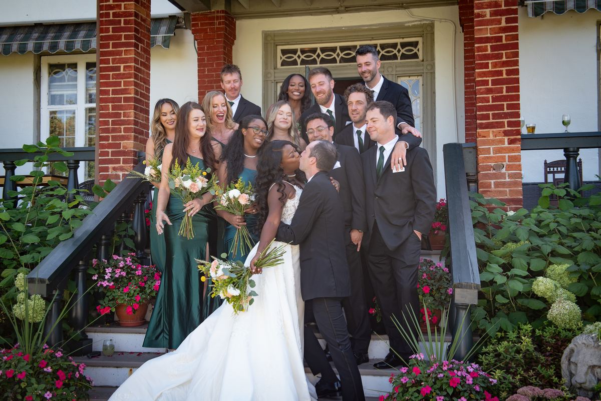 Bride and groom kiss amid laughing bridesmaids in green gowns and groomsmen in black suits, surrounded by vibrant flowers.