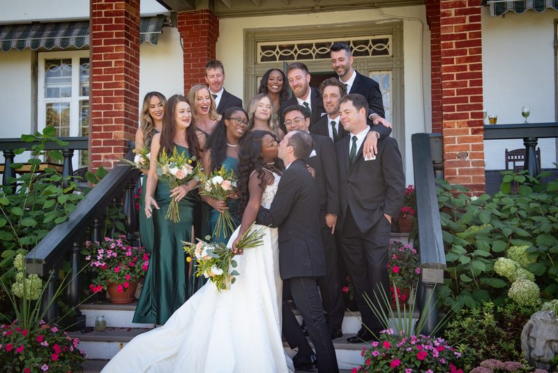 Bride and groom kiss amid laughing bridesmaids in green gowns and groomsmen in black suits, surrounded by vibrant flowers.