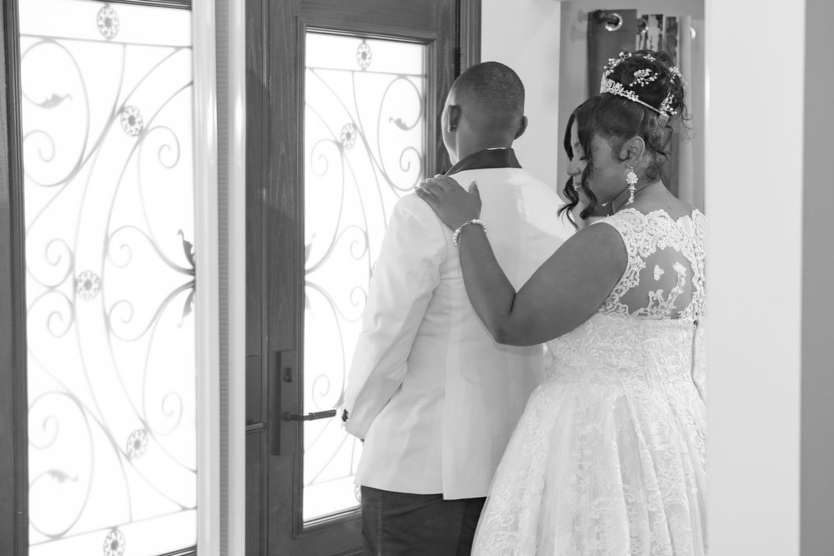Bride and groom stand back-to-back, gazing through an ornate glass door, her hand resting tenderly on his shoulder.