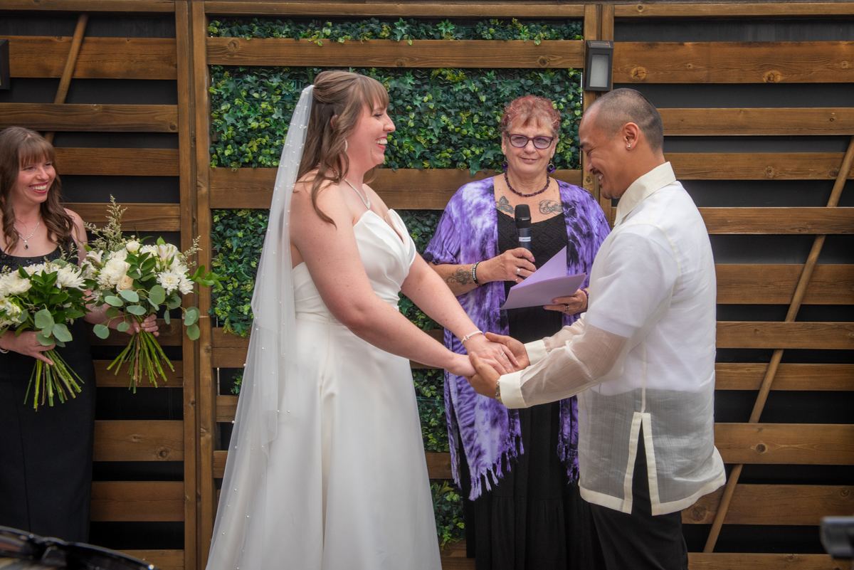Bride and groom hold hands smiling during ceremony with wooden backdrop and greenery, blending Western and Filipino attire.