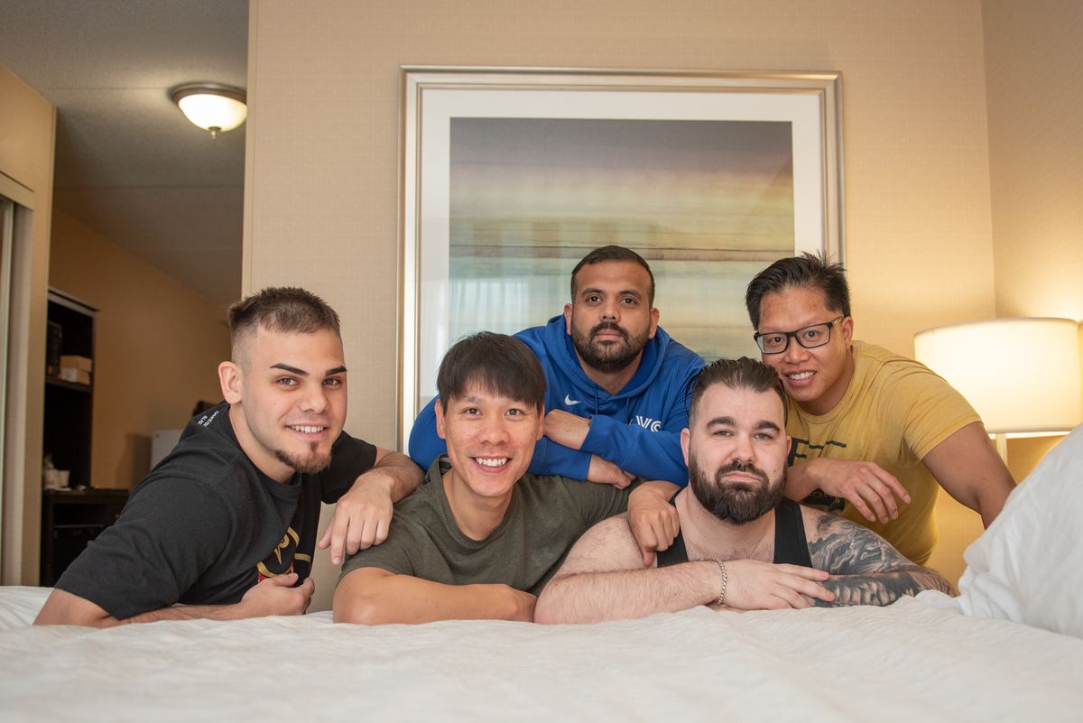 Five young men pose joyfully on a bed in a warmly lit room, their relaxed postures and smiles conveying camaraderie.