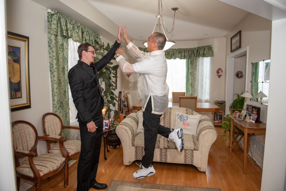 Two men in high-fives in a cozy living room with floral curtains and vintage chairs, illuminated by soft light.