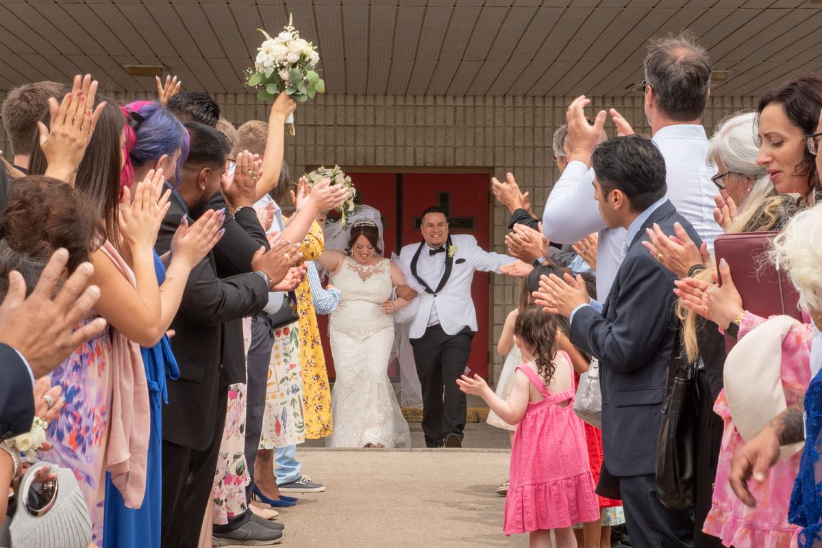A smiling bride and groom exit a church with guests cheering and waving, creating a joyful corridor.