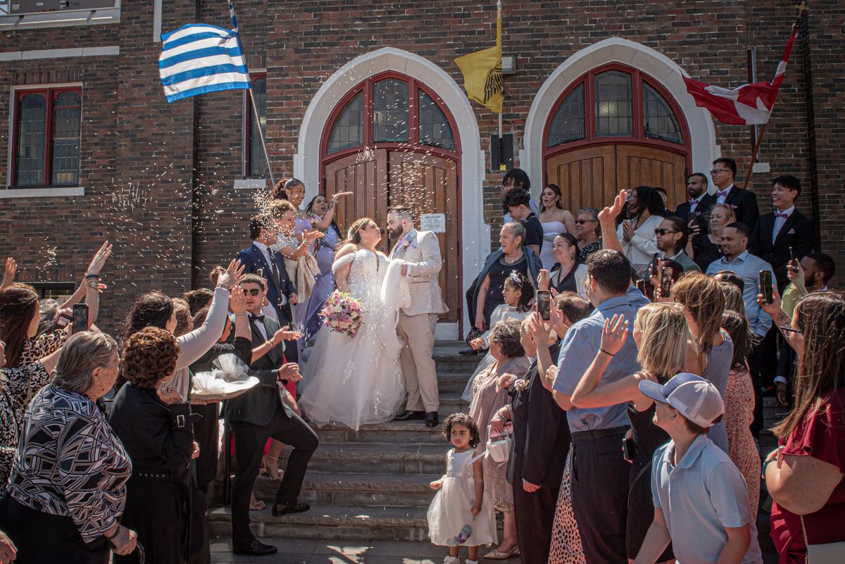 A bride and groom exit a church, showered in confetti, kissing as guests cheer with raised phones and flags of Greece.