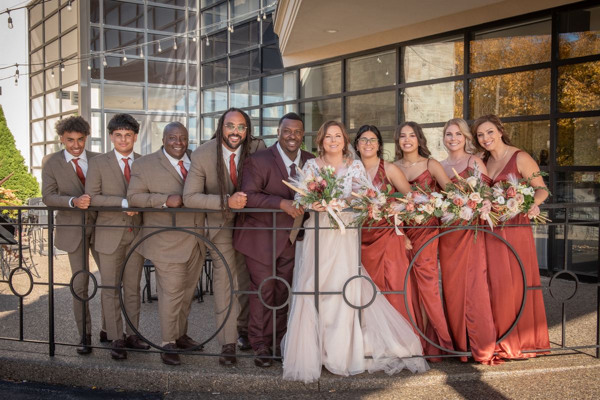 A bride and groom with their wedding party on a sunny balcony, surrounded by autumn foliage and modern architecture.