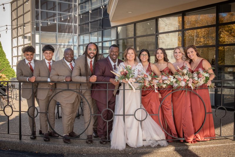 A bride and groom with their wedding party on a sunny balcony, surrounded by autumn foliage and modern architecture.