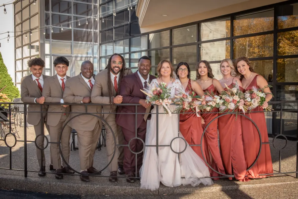 A bride and groom with their wedding party on a sunny balcony, surrounded by autumn foliage and modern architecture.