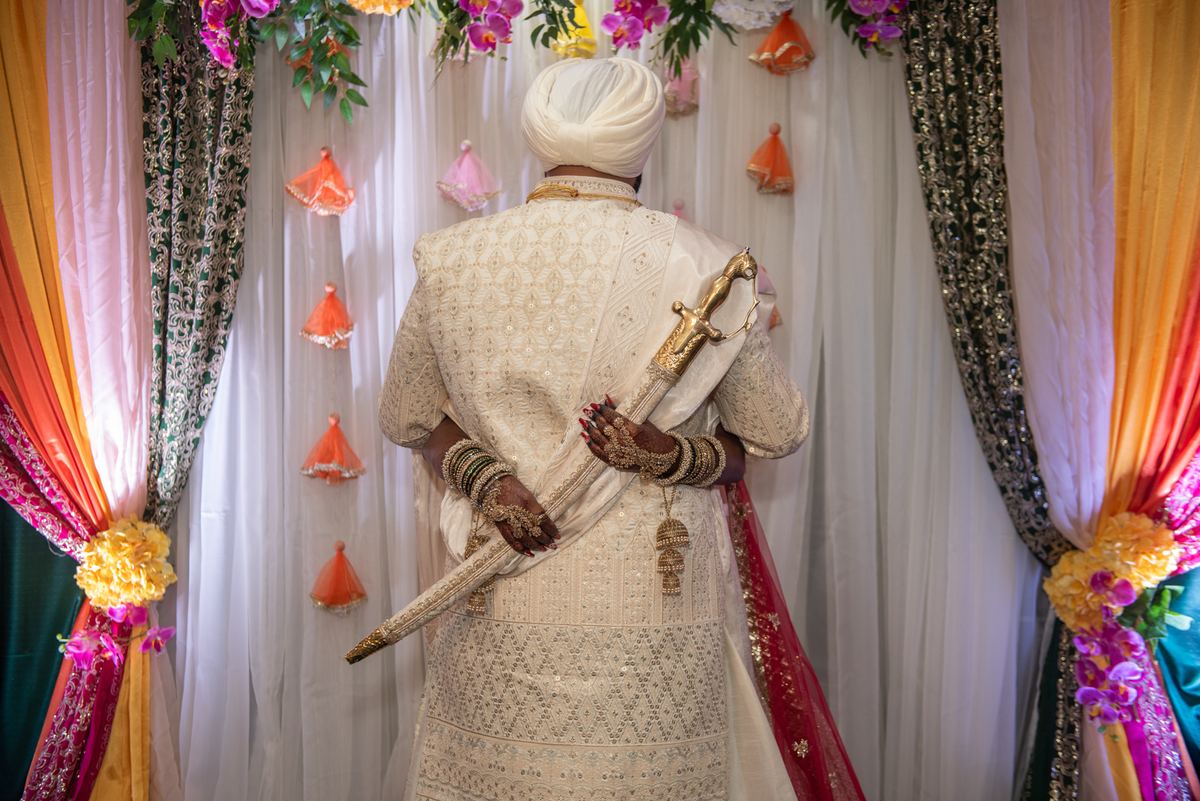 A man in ornate attire holds a ceremonial sword at a festive celebration with draped fabrics and floral tassels.