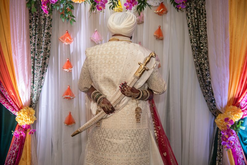 A man in ornate attire holds a ceremonial sword at a festive celebration with draped fabrics and floral tassels.