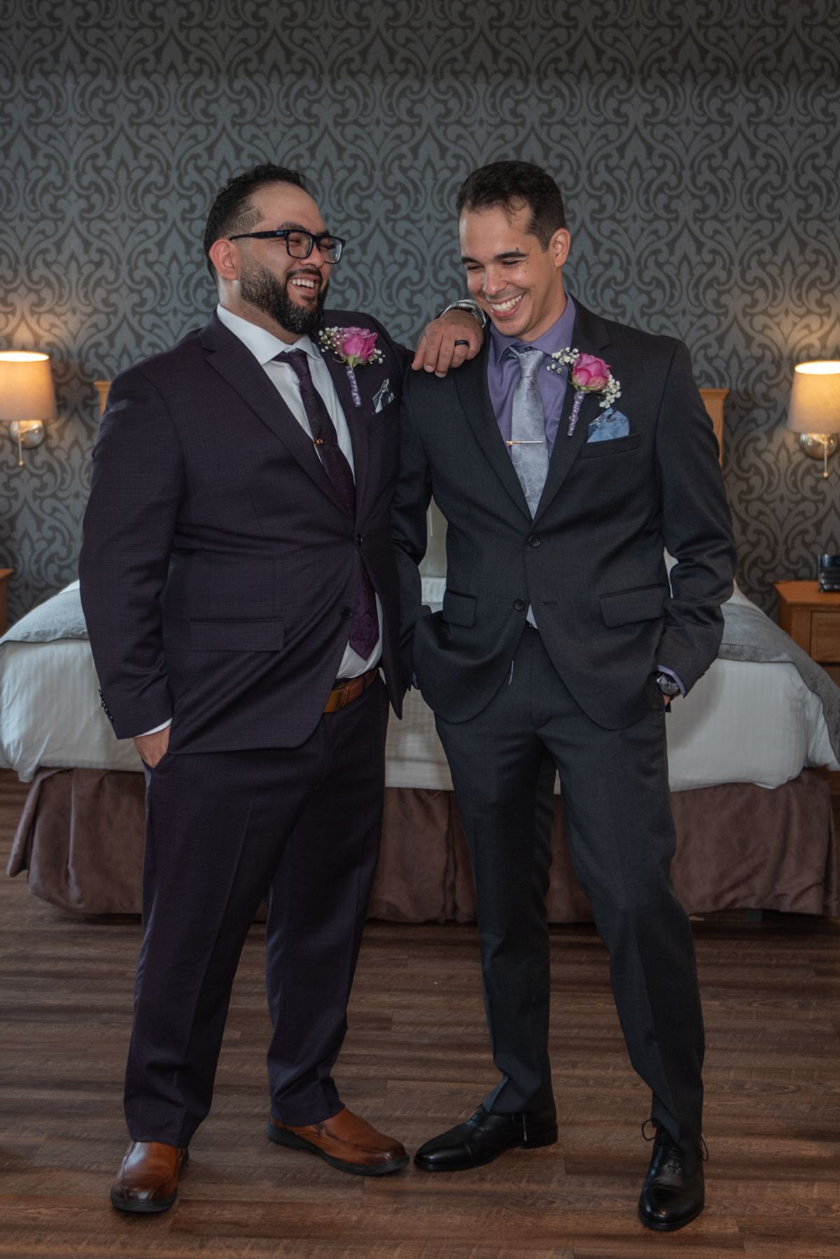 Two men in formal wedding attire laugh joyfully in a warmly lit hotel room, one arm around the other.