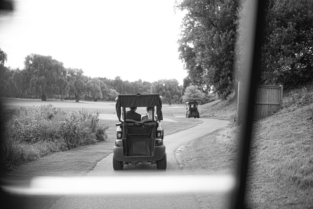 A couple rides in a golf cart along a course, surrounded by foliage and a wooden fence on an overcast day.