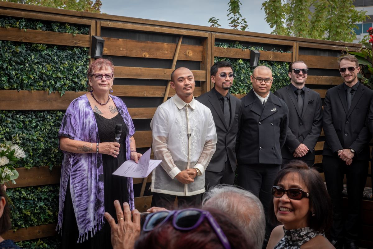 A woman in a purple shawl speaks into a microphone at an outdoor gathering with formal attire and greenery.