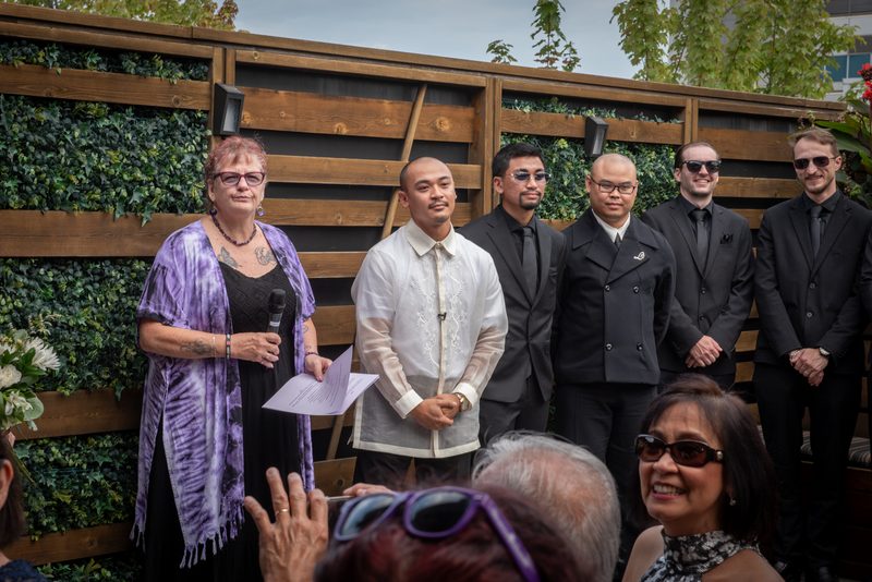 A woman in a purple shawl speaks into a microphone at an outdoor gathering with formal attire and greenery.