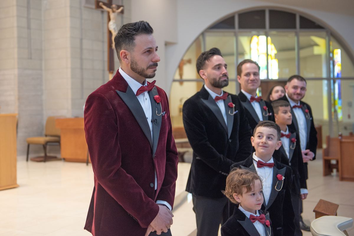 A groom and his party in formal red attire stand solemnly in a church with soft light from stained glass windows.