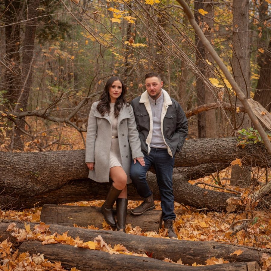 A couple sit together on fallen logs in a wood of bare trees and deep autumn leaf litter.
