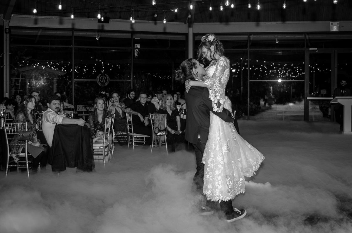 Bride and groom dance above foggy floor, guests watching from background, twinkle lights casting glow.