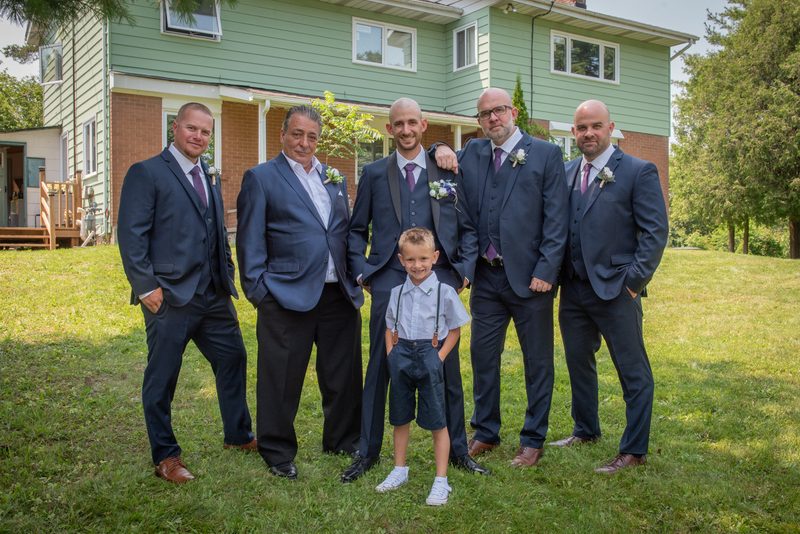 A wedding party poses on a lawn in front of a house, the smiling groom with his groomsmen and ring bearer.