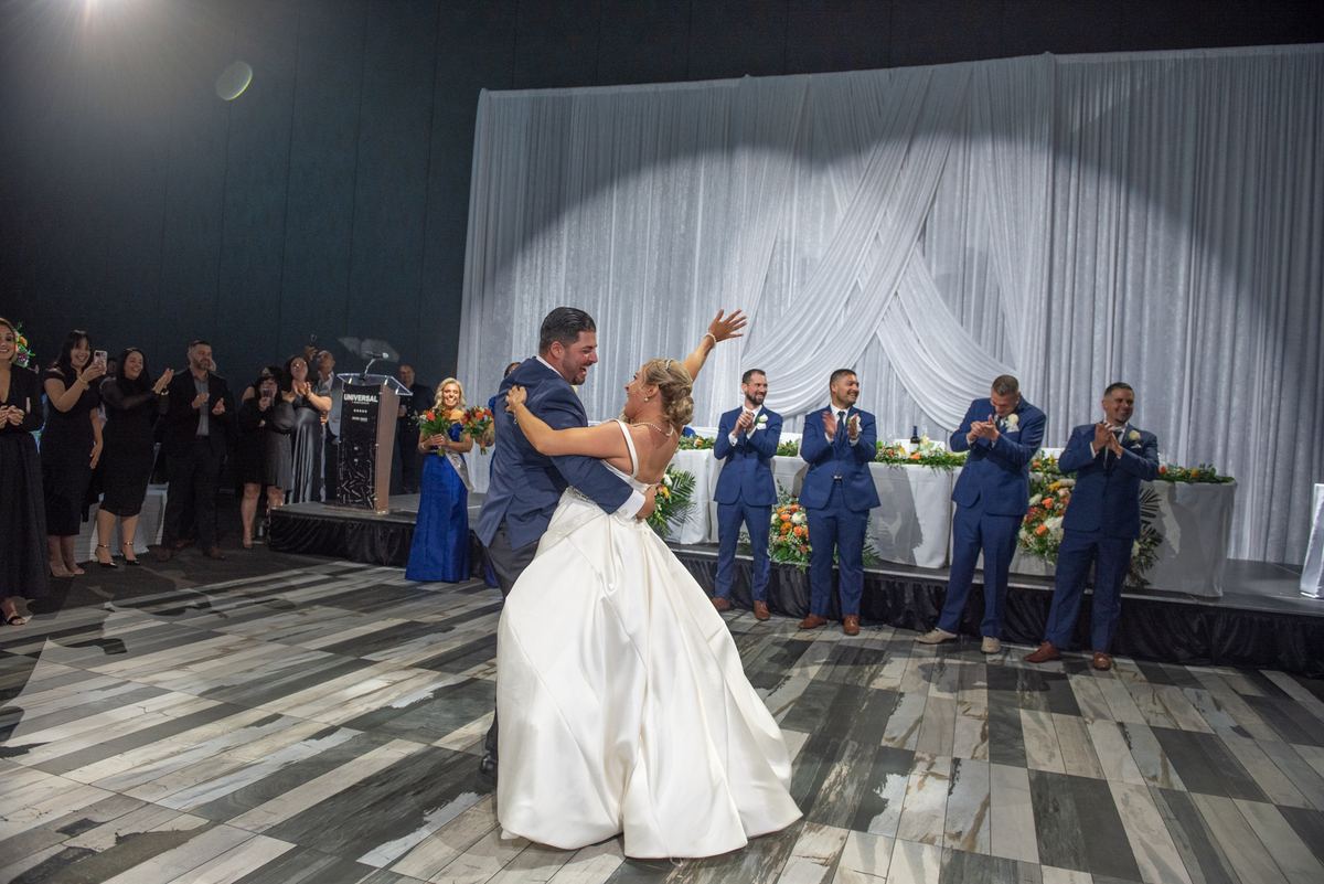 Bride and groom dance center stage under spotlight's circular glow, guests in formal attire clap along edges.