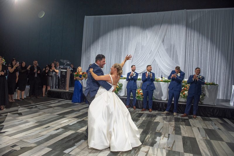 Bride and groom dance center stage under spotlight's circular glow, guests in formal attire clap along edges.