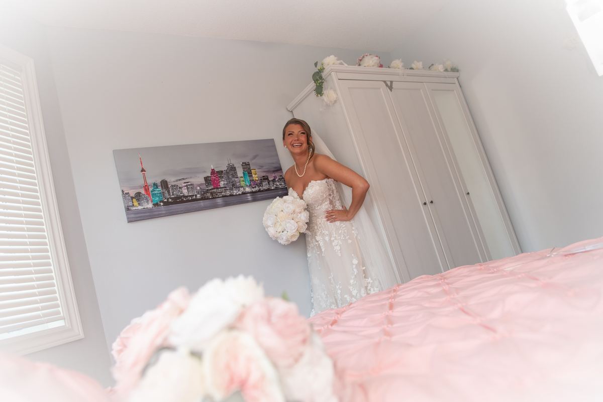 A bride smiles and holds a bouquet near a wardrobe in her bedroom, with floral arrangements on the bed.