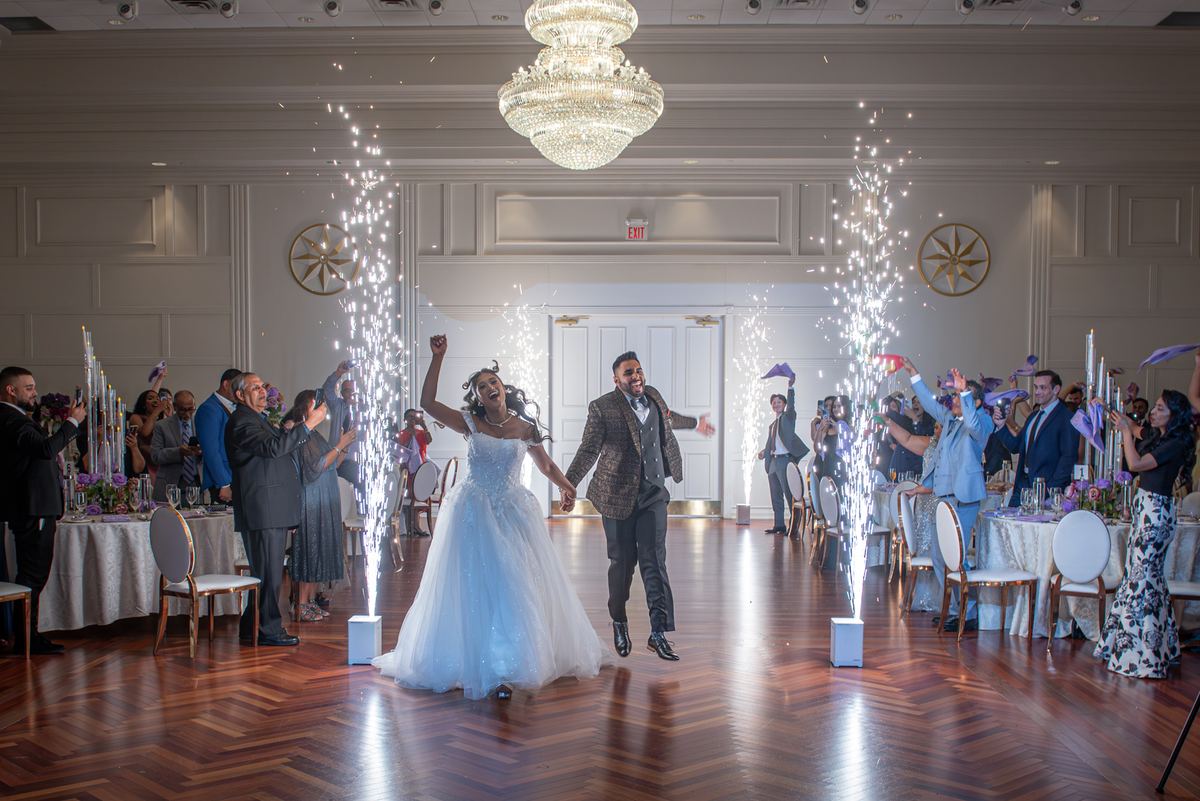Bride and groom run down the center of a ballroom, arms raised, as guests cheer with purple decorations.