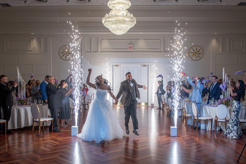 Bride and groom run down the center of a ballroom, arms raised, as guests cheer with purple decorations.