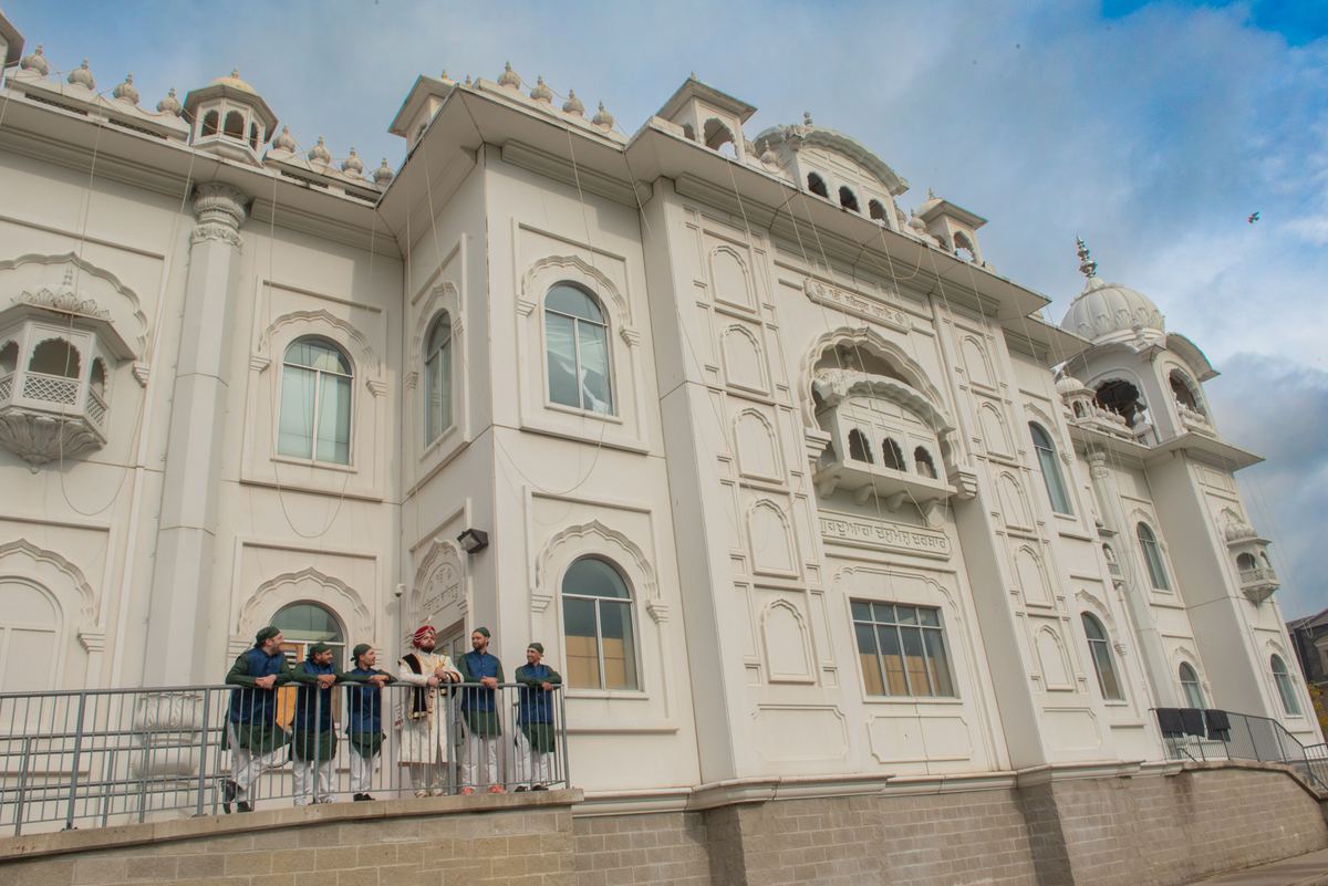Men in Sikh attire stand on a balcony railing before a grand white gurdwara with ornate domes, under a partly cloudy sky.