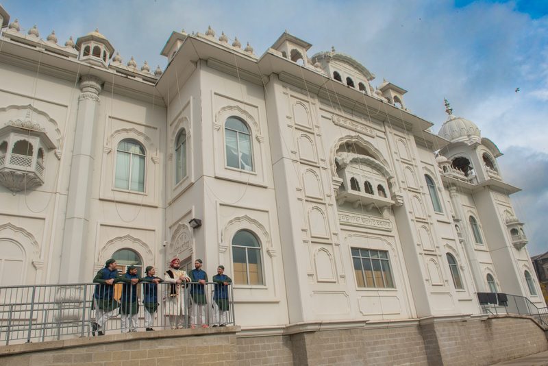 Men in Sikh attire stand on a balcony railing before a grand white gurdwara with ornate domes, under a partly cloudy sky.