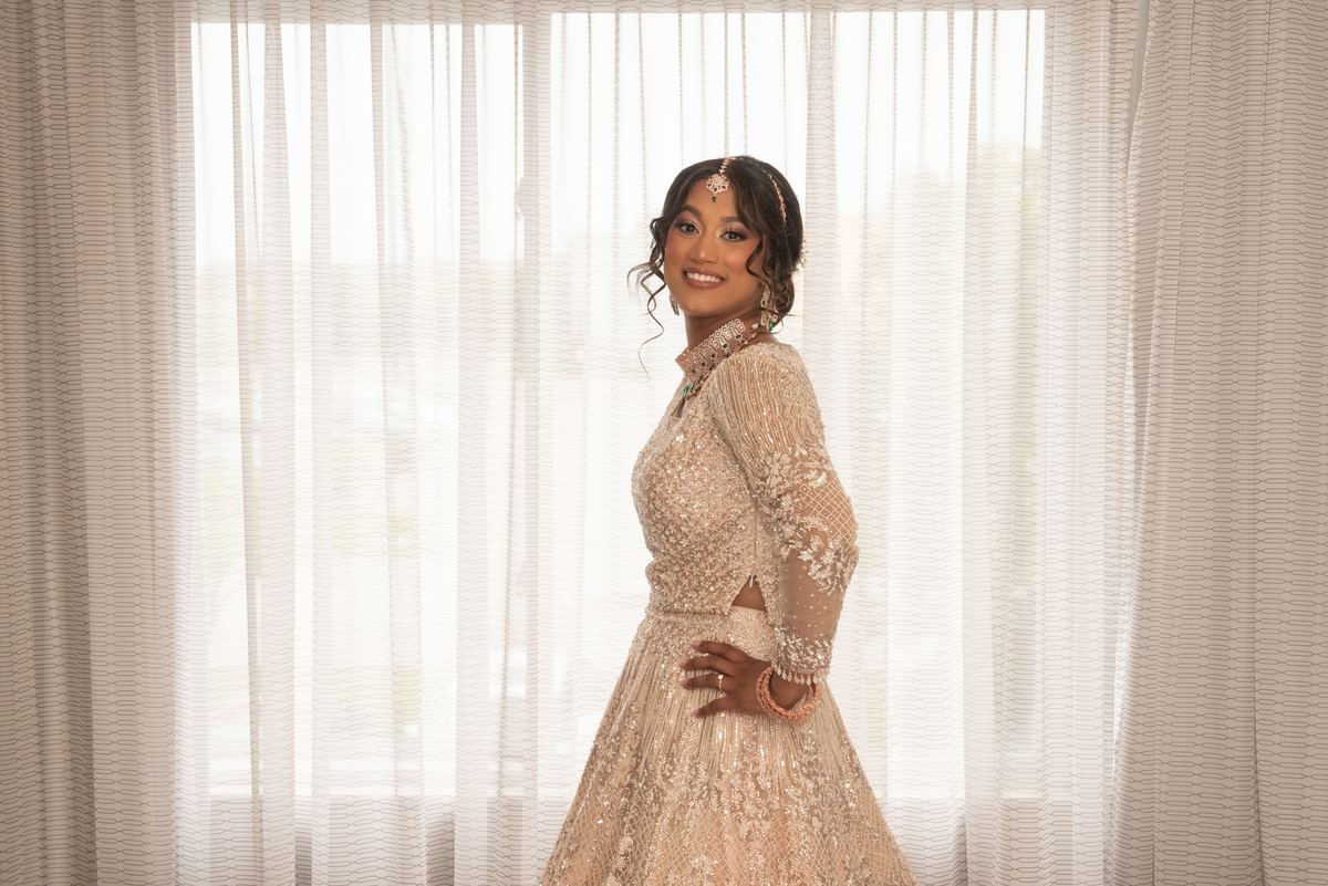 A smiling bride in an ornate lehenga stands before a sheer curtain, adorned with traditional jewelry.