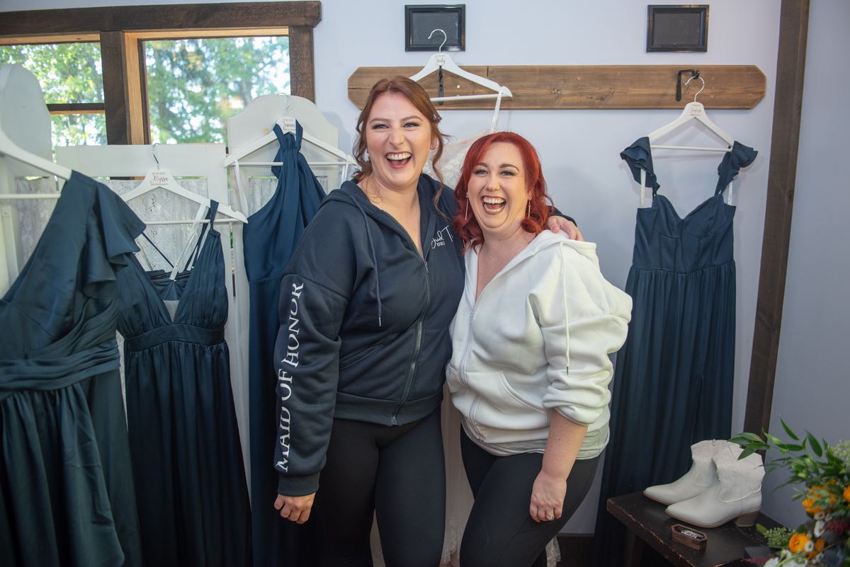 Two women in dark teal bridesmaid dresses laugh together in a shop with racks of gowns, one wearing a Maid of Honor hoodie.
