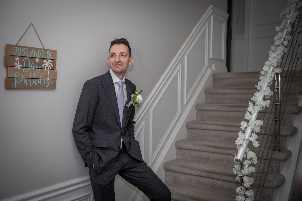 A groom in a dark suit and light lavender tie leans against a staircase adorned with white floral garlands, smiling upward.