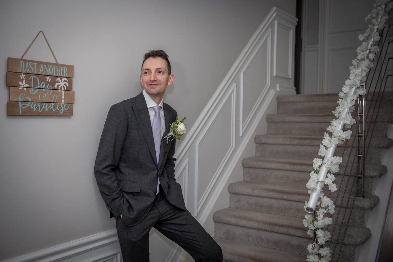 A groom in a dark suit and light lavender tie leans against a staircase adorned with white floral garlands, smiling upward.