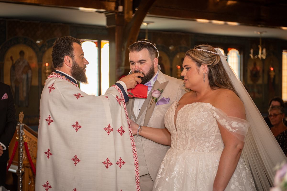 Bride and groom stand beside priest, sharing a chalice during wedding ceremony in warmly lit church with stained glass light.