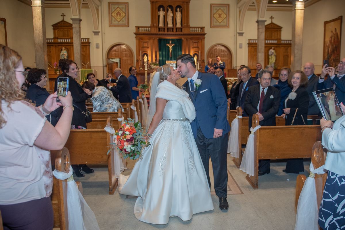A bride and groom kiss while walking down the aisle in a warmly lit church, surrounded by guests taking photos.